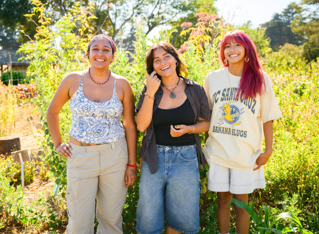 Three Future Organic Farmers grantees smiling together in a vibrant organic garden, representing the next generation of organic farmers
